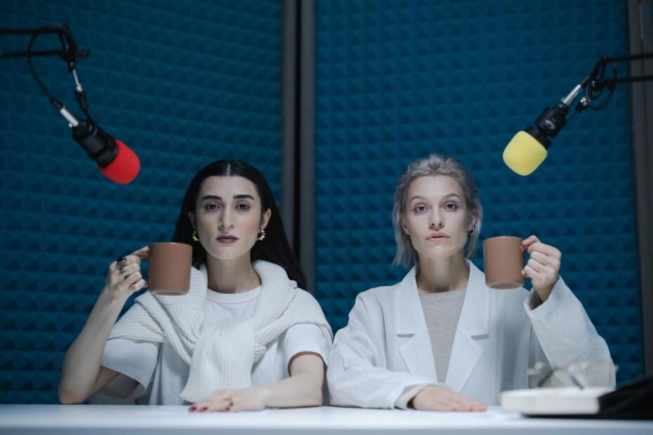 Two women seated together with microphones, podcasting in a studio holding cups